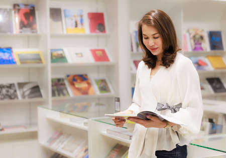 Portrait of Asain pretty cheerful female cozy standing and reading book in library and leaning on glass wall with happy, focus and concentrate on contents.の写真素材