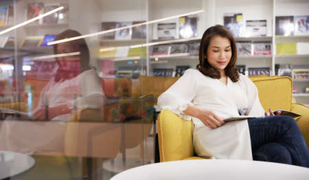 Portrait of Asain pretty cheerful female cozy sitting on sofa and reading a book in library with calm and happy. Mature woman focus and concentrate on content, self-education concepts.の写真素材
