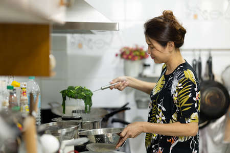 An authentic real shot of Asian woman boiling and cooking green vegetables for food in kitchen. Housewife prepares healthy dinner for her family with happy face.の写真素材