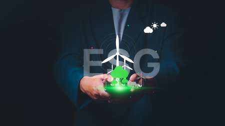 Businessman holding a smartphone with a glowing ESG icon, wind turbines, solar panels, and green...