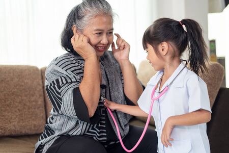 Little cute Asian granddaughter wearing a nurse uniform with stethoscope examines an elderly grandmotherの写真素材