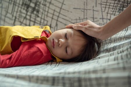 Sick Asian little girl lying on sofa while her mother is touching her forehead
