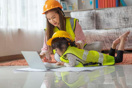 Little Asian child engineer in yellow helmet using laptop with her mother, education and occupation...
