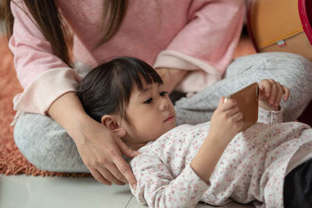 Mother and her little daughter enjoy leisure time using a smartphone at home, Happy Asian child, family conceptの写真素材
