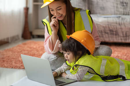 Little Asian child engineer in yellow helmet using laptop with her mother, education and occupation conceptの写真素材
