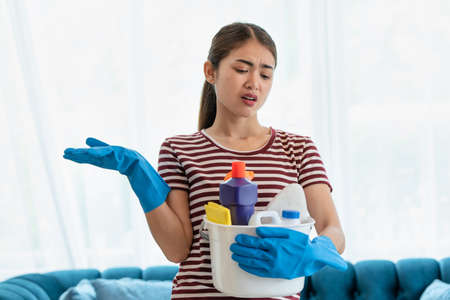 Stressed young Asian housewife wearing gloves and holding basket with cleaners and brushes feeling tired and frustrated, cleaning and washing conceptの写真素材