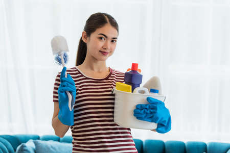 Happy young Asian housewife wearing gloves and holding basket with cleaners and brushes, cleaning and washing conceptの写真素材