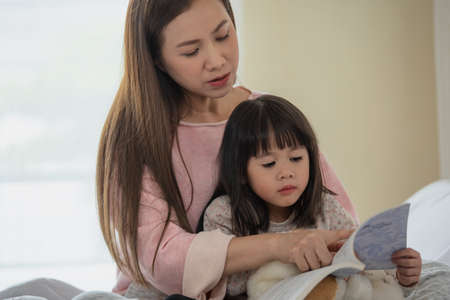 Happy family mother and child daughter read a book on bed in bedroom, Asian family loving conceptの写真素材
