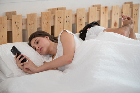 Young couple in bed using mobile phone lying backs to each other, relationship and communication problems, internet social network addictionの写真素材