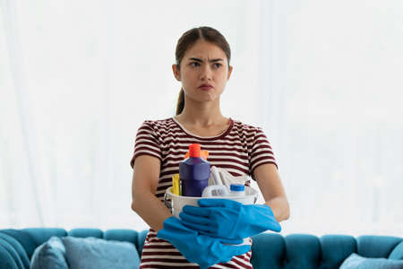 Stressed young Asian housewife wearing gloves and holding basket with cleaners and brushes feeling...