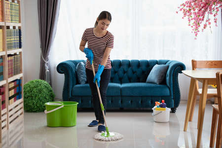 Happy young Asian housewife wearing gloves washing floor with mop in living room, Cleaning serviceの写真素材