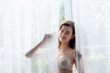 Portrait of Sexy Asian woman opening the curtain after waking up in the morningの写真素材