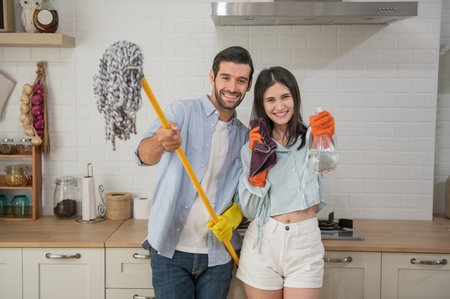 Happy young couple cleaning up in the kitchen, they hold a mops and rags with spray bottle in hands, Happy family conceptの写真素材