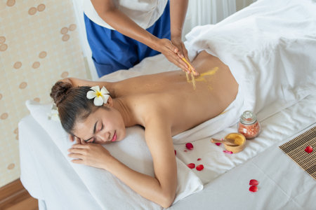 Young Asian woman enjoying in spa salon, Beauty therapist pouring scrub on woman back at health spa, Scrubbing and skin care conceptの写真素材