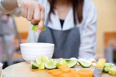 Asian Thai people holds and squeezes green lime (lemon) on the top of white little bowl with quater limes on wood plate. The environment is simple wood kitchen theme.の写真素材