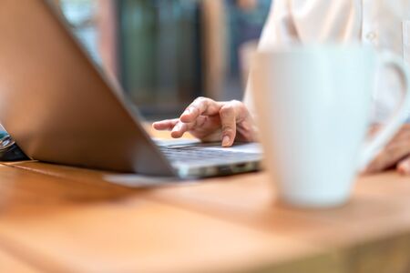 Close up asian man's hand and finger work on his laptop with coffee cup foreground and wood...