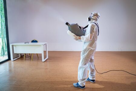 Professional technical man in prevention suit sprays sterilising solution by electrical spray machine on wood floor and white background with studio light.の写真素材
