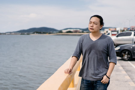 Asian Male long hair with dark gray t-shirt stands on the bride beside the road and looks forward to the sea in the afternoon period.の写真素材