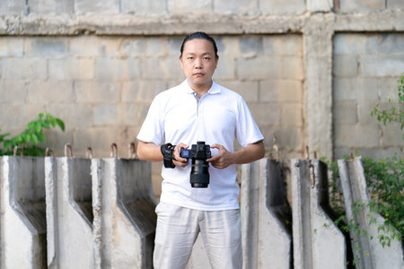 Asian man serious looks at the camera while holds th mirrorless camera medium format in the hand on the construction concret wall background.の素材 [FY310167338638]