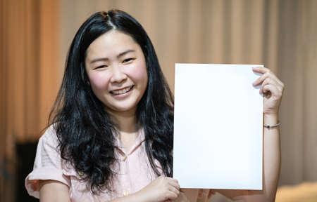 The Asian woman is holding and showing empty white A4 paper portrait - vertical angle on orange brown background.の写真素材