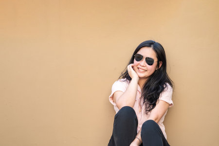 Relaxed Pretty Asian Woman sits on the rooftop floor with a light brown background and smiles to the camera.の写真素材