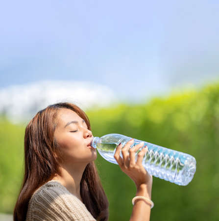 Asian Woman drinks big bottle water in outdoor green bokeh tree background.