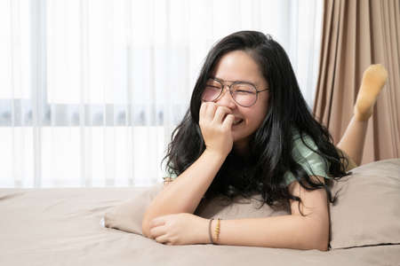 Beautiful glasses Asian woman feels embarrassed and smiles to the camera and lays down on her bed ...