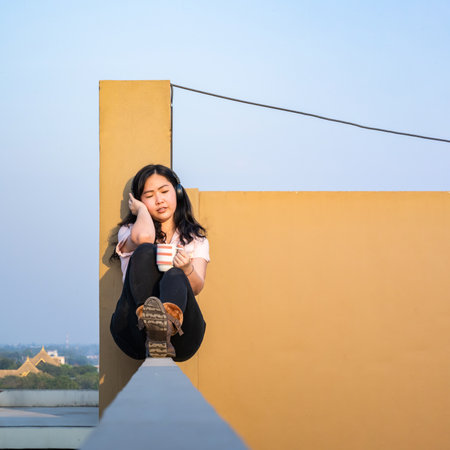 Glasses Asian woman is listening music via headset, drinking coffee on rooftop barricade cement (not dangerous because the opposite site is rooftop floor as well).の写真素材