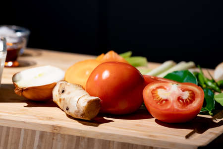 Tomato and sliced half of tomato beside it with onion and galangal, on wood plate in studio light...