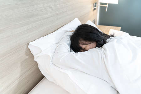 Asian Lady in white long arm t-shirt pajamas lie prone, lie on the stomach in bed with tried and exhausted mood and tone.の写真素材