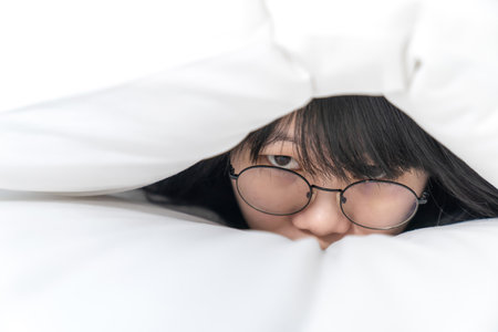 close up to the Cute Asian Glasses woman, she hides her body inside white pillow on the bedの写真素材