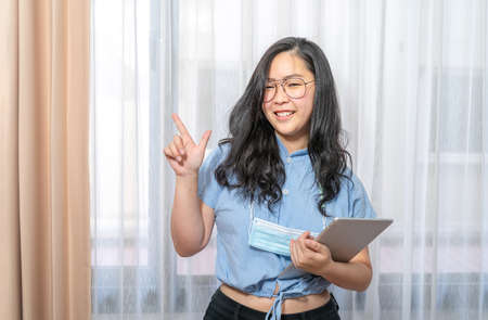 Happy Asian woman fingering up and pointing with tablet computer on her hand at copy space while...