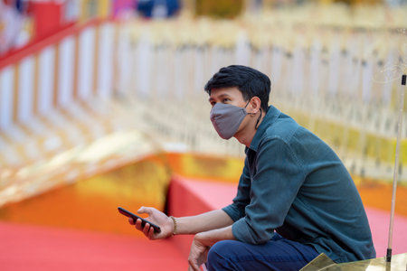 Asian Smart man in casual dark green shirt and gray cloth mask sit in the outdoor red carpet step and hold the mobile phone on his hand.の写真素材