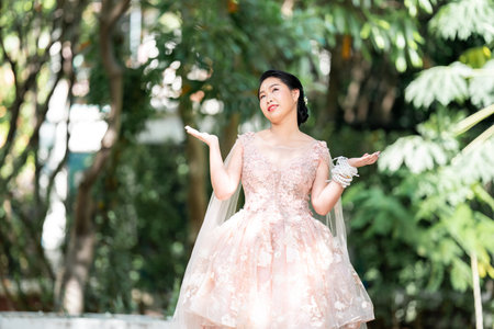 Asian girl in an old rose pink color knee length fluffy dress with high heel slingback shoes and ceremonial thread on her wrist. She is brighter mood  posing in the shade of tree garden.の写真素材