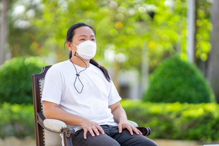 A man wearing white T shirt and white mask is sitting calm on the armchair with his arms on the armrest and his back on the backrest.の写真素材