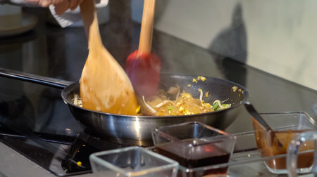 A cook stirs fry Pad Thai noodles in a pan with spatulas on electric stoves. Sauce dishes for...