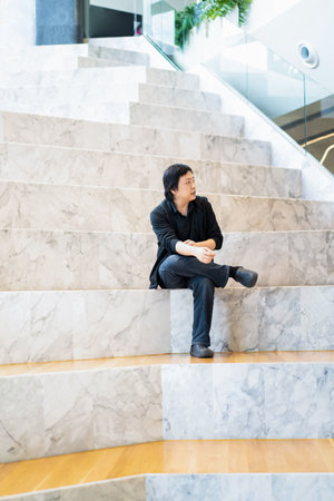 Asian Man in Black cardigan and polo shirt sits on the white grand marble stairs in the grandstand with wood floor on its step.の写真素材
