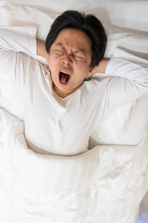 An Asian man is captured in a moment of genuine yawn as he rests on a soft white bed and pillow. The image evokes a sense of contagious yawning, inviting viewers to join in the action.の写真素材