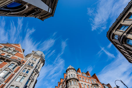 A bottom-up view from the middle of a busy intersection, looking up at the four surrounding buildings against a clear blue sky with a few scattered clouds in London, England.の写真素材