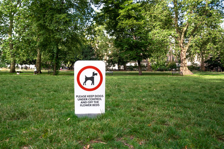 Sign in a park asking to control dogs and keep them off flower beds. Surrounded by green grass trees and public space.の写真素材