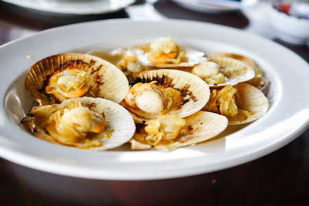 Grilled scallops in the shell with butter, a fresh seafood menu of a restaurant, served on a white plate on the wooden table with Thai seafood sauce.の写真素材