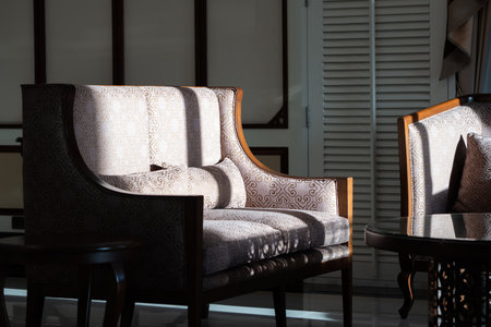 Sunlight shines on the elegant wooden sofa upholstered with ivory velvet fabric and a round coffee...
