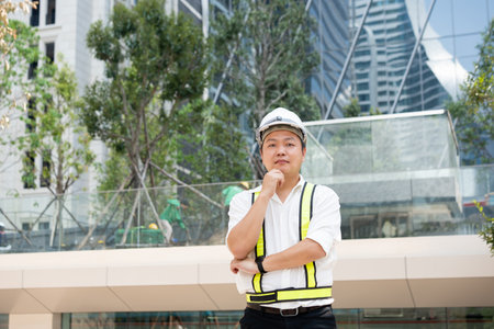 A confident Asian construction engineer wearing a white safety helmet and reflective vest, standing with arms crossed and one hand on his chin, smiling slightly with confidence.の写真素材