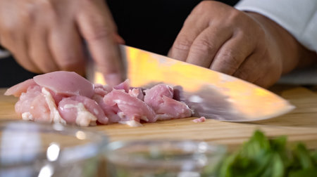 A chef using a kitchen knife slices uncooked chicken meat on a chopping board, the close up view of preparing food ingredients.の写真素材