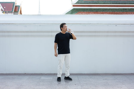 A tourist drinks water in front of a plain white temple wall on a hot day, taking a short break while exploring Thai culture.の写真素材