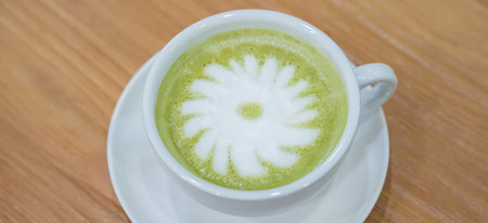 A warm cup of matcha latte rests on a white saucer atop a wooden table, with white foam forming a...