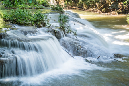 A small waterfall cascades over layered rocks surrounded by trees, with the water flowing in shallow steps into a wider pool.の写真素材