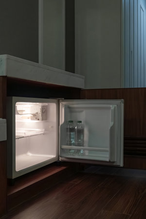 A compact fridge is built under the counter with a wooden door open beside it. The calm room lighting adds a soft tone to the interior setup.の写真素材