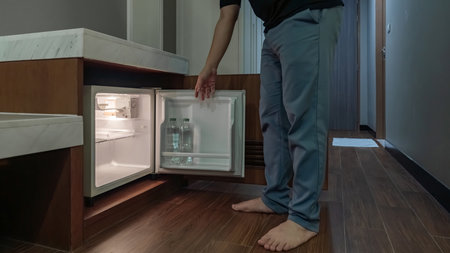 A man opens a small fridge built into a wooden cabinet in a dim hotel room, and the warm interior light contrasts with the dark surroundings.の写真素材