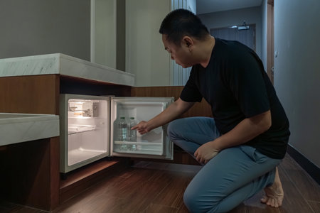 A man opens a small fridge built into a wooden cabinet in a dim hotel room, and the warm interior light contrasts with the dark surroundings.の写真素材